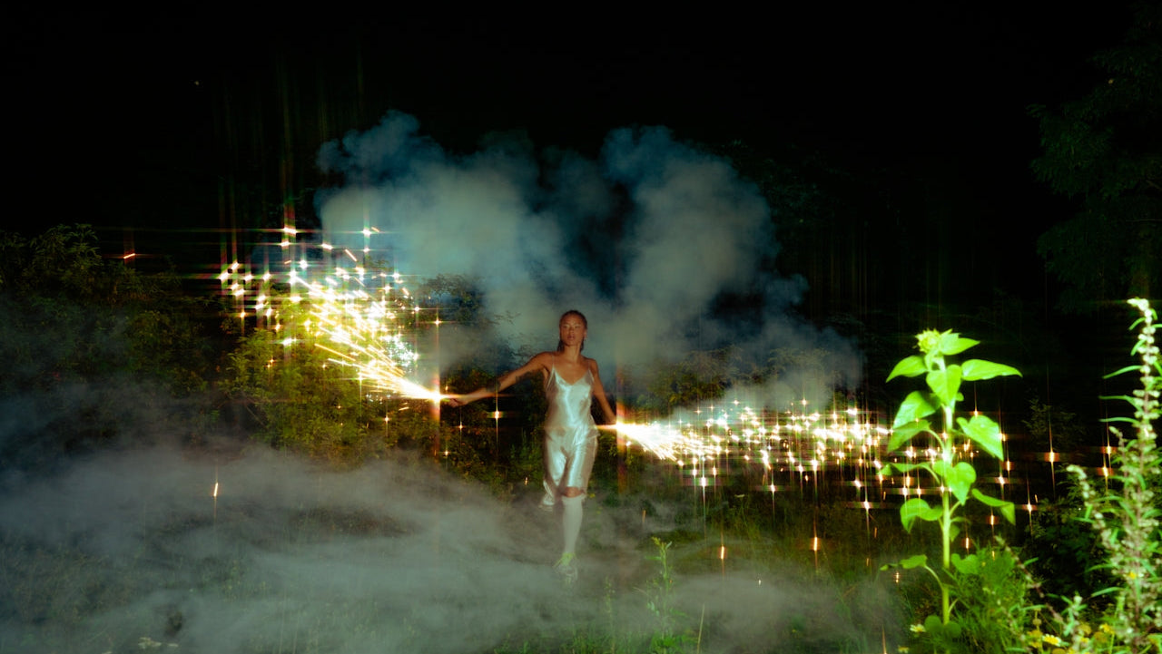 Person holding sparklers in a dark outdoor setting with smoke and lights.